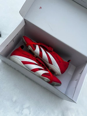 Adidas Predator röd/vit fotbollssko för junior - Säljer ett par Adidas Predator fotbollsskor i rött och vitt med klassiska tre ränder på sidan och texten 'PREDATOR' under sulan. Skorna har snörning och är designade för gräsplan. Snygg och sportig look som sticker ut på planen.