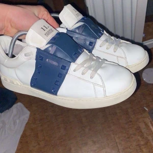 Valentino Rockstud  - Snygga Valentino Rockstud Open sneakers i vitt skinn med bred blå rem över vristen dekorerad med ikoniska nitar. Klassisk rund tå, platt sula och vita skosnören. Kommer med originalkartong, dustbag och tags. Perfekta för dig som vill sticka ut med lyxig streetstyle. 2-3 nitar saknas och även lite slitage där framme men syns inte så mycket 