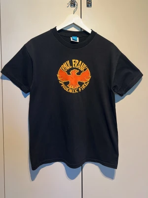 Svart Paul Frank Phoenix Fire t-shirt - Cool svart t-shirt från Paul Frank med färgglatt tryck av en orange och gul fågel samt texten 'Phoenix Fire' på bröstet. Klassisk passform och rund halsringning, perfekt för dig som gillar retro vibes och statement-plagg.