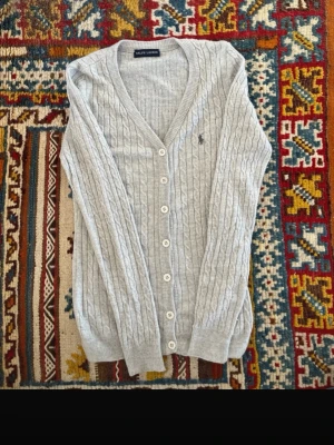 Grå kabelstickad kofta från Ralph Lauren - Snygg grå kabelstickad kofta från Ralph Lauren med klassisk v-ringning och knäppning framtill. Koftan har långa ärmar, ribbade muddar och den ikoniska broderade loggan på bröstet. Perfekt för lager på lager och en stilren look.