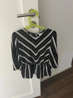 Randig svartvit topp med volang - Snygg topp med svart botten och vita samt ljusrosa ränder. Modellen har trekvartsärmar och volang nedtill som ger en trendig siluett. Mjuk och lätt material, perfekt till jeans eller kjol. Rund halsringning och coolt mönster med diagonala ränder.