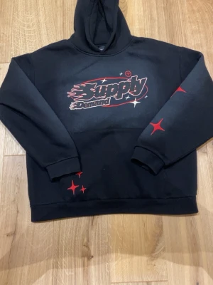 Svart hoodie Supply & Demand - Svart hoodie från Supply & Demand med stor logga i vitt och rött framtill. Hoodien har röda stjärndetaljer på ena ärmen och nedtill. Klassisk huva och ribbade muddar. Perfekt för en streetwear-look.