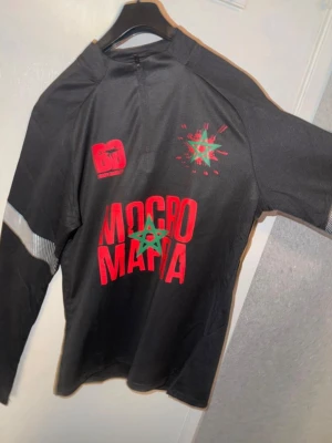 Svart långärmad tröja Mocro Mafia - Svart långärmad tröja från Mogro Mafia med röd och grön tryckt logga och text framtill. Tröjan har silvriga detaljer på ärmarna och en sportig vibe. Materialet känns som polyester eller syntet, perfekt för dig som gillar streetwear.