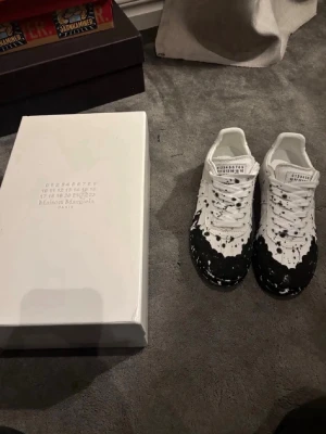 Maison Margiela vita sneakers med svart färgstänk - Maison Margiela sneakers i vitt läder med unika svarta färgstänk över tån och sidorna. Klassisk låg modell med vita snören och märkets ikoniska etikett på plösen. Snygg och konstnärlig design som sticker ut i mängden.