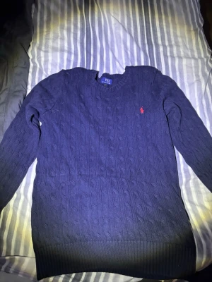 Mörkblå kabelstickad tröja Polo Ralph Lauren - Kabelstickad mörkblå tröja från Polo Ralph Lauren i bomull. Klassisk rund halsringning och ribbade muddar. Tröjan har det ikoniska röda Polo-märket broderat på bröstet. Perfekt för dig som gillar stilrena och tidlösa plagg. NYPRIS 2100kr