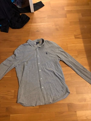 Grå skjorta från Ralph Lauren - Snygg grå skjorta från Ralph Lauren med klassisk krage och knappar framtill. Skjortan har lång ärm och den ikoniska loggan broderad på bröstet. Tillverkad i mjuk bomull som känns lätt och bekväm. Perfekt för en clean och stilren look.