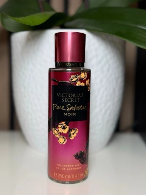 Victoria's Secret Pure Seduction Noir - En snygg parfymmist från Victoria's Secret i doften Pure Seduction Noir. Flaskan är metallicrosa med svarta och guldiga detaljer samt blommotiv. Rymmer 250 ml och har en elegant, rund form med matchande lock.