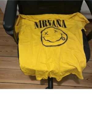 Gul Nirvana t-shirt med tryck - Gul t-shirt med stort svart Nirvana-tryck och det klassiska smiley-ansiktet på framsidan. Perfekt för dig som gillar bandmerch och vill sticka ut med en färgstark look.