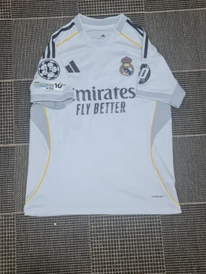 Real Madrid matchtröja Adidas vit - Säljer en vit Real Madrid matchtröja från Adidas med klubbmärke och sponsortryck på bröstet. Tröjan har svarta detaljer, gula linjer på sidorna och UEFA Foundation-märke på ärmen. Tillverkad i lätt och ventilerande material (AEROREADY). Perfekt för fotbollsträning eller support.