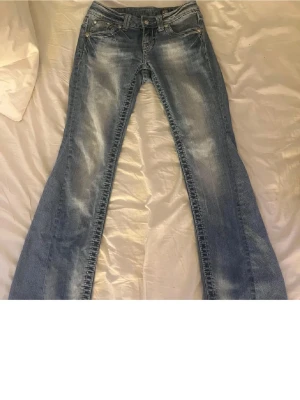 Blå bootcut jeans från Miss Me - Säljer ett par blå bootcut jeans från Miss Me med snygg tvätt. Jeansen har dekorativa nitar på fickorna och klassisk femficksmodell. Tillverkade i denim med en cool vintagekänsla och markerade sömmar. Byxorna är före detta skinny men professionellt omsydda till bootcut