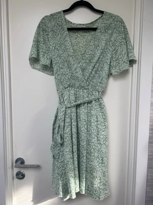 Grön blommig omlottklänning - Söt grön kortklänning med vitt blommönster och omlottmodell. Klänningen har korta ärmar, v-ringning och ett matchande knytband i midjan. Perfekt för varma dagar och ger en fräsch sommarkänsla.