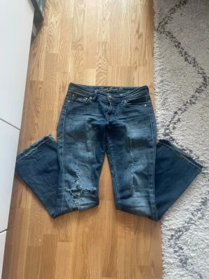 Blå slitna bootcut jeans - Säljer ett par blå bootcut jeans med slitningar och hål på ena benet. Jeansen har klassisk femficksdesign, låg midja och är gjorda i denim. Perfekta för en avslappnad och trendig look.