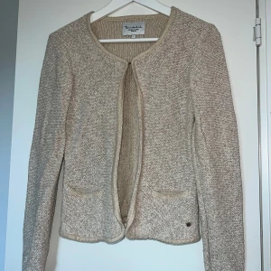 Beige stickad kofta från Bondelid - Snygg beige stickad kofta från Bondelid med öppen front och rundad halsringning. Koftan har två små fickor framtill och ribbade detaljer vid kanterna. Perfekt för lager på lager och enkel att matcha med olika outfits.