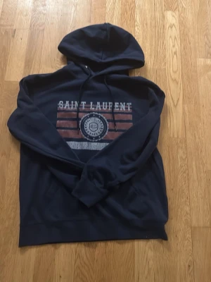 Mörkblå Saint Laurent hoodie - STORLEK M. Mörkblå hoodie från Saint Laurent med tryck framtill i vitt och rött samt emblem. Klassisk huva med dragsko och långa ärmar. Perfekt för en avslappnad och cool stil. Mjuk bomullskänsla och ribbade muddar vid ärmslut och nederkant. Använd en gång