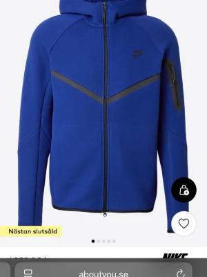 Blå Nike hoodie med dragkedja - Snygg blå hoodie från Nike med hel dragkedja framtill och svarta detaljer. Hoodien har en huva, svarta ränder över bröstet och en praktisk ficka med dragkedja på ärmen. Perfekt för en sportig och avslappnad stil.