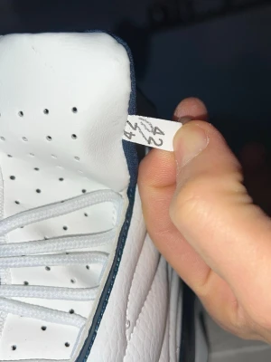 Vita sneakers med blå detaljer - Snygga vita sneakers med mörkblå detaljer och texten 'SPORTSPORT' på plösen. Skorna har perforerad ovandel och vita skosnören. Röd logga framtill och vadderad insida för extra komfort. Perfekta för dig som gillar sportig stil.