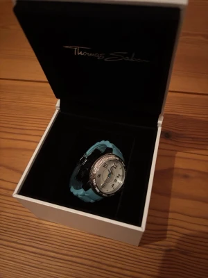 Silverfärgad barnklocka Thomas Sabo - Snygg klocka från Thomas Sabo med silverfärgat ur och ljusblått silikonarmband. Uret har glittrande stenar runt boetten och tydliga siffror på vit urtavla. Väldigt gammal men knappt använd. 