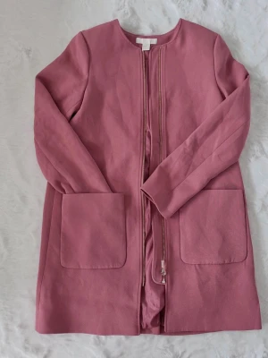 H&M gammelrosa kavaj/kappa strl 32 - H&M gammelrosa kavaj/ kappa strl 32  En jättefin, stilren kappa i en härlig dov rosa nyans (gammelrosa). Kappan har en modern, kraglös design och en praktisk dragkedja framtill, vilket ger ett stilrent och minimalistiskt intryck. Detaljer:  * Märke: H&M  * Färg: Gammelrosa / Mörkt puderrosa  * Modell: Rak passform, kraglös med guldfärgad dragkedja  * Fickor: Två rymliga utanpåfickor framtill  * Skick: Mycket sparsamt använd