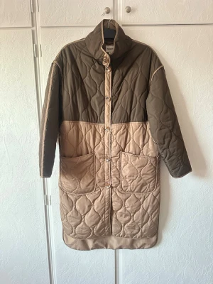 Quiltad lång kappa i khakigrönt och beige st S - Snygg quiltad kappa från Kappahl. Märkt st S och är rymlig i storlek. Finns ett litet litet märke, se bild, annars jättefin 