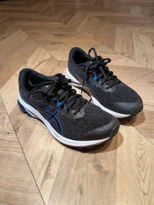 Svarta Asics sneakers med blå detaljer - Svarta sneakers från Asics med blå detaljer. Ovandel i mesh för bra andningsförmåga, svarta skosnören och vit sula med svarta och blå inslag. Sportig design som passar till streetwear och träning.