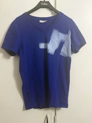 Blå t-shirt med grafiskt tryck från  J.Lindeberg - Snygg blå t-shirt från   J.Lindeberg med v-ringning och ett vitt grafiskt tryck över bröstet. T-shirten har korta ärmar och är gjord i mjuk bomull, perfekt för en avslappnad och trendig stil.