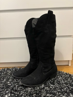Svarta cowboy boots i mocka - Snygga svarta cowboy boots i mjuk mocka med klassisk spetsig tå och låg klack. Höga skaft med dragflikar upptill för enkel påtagning. Perfekta för dig som vill ha en stilren westernlook. Skorna är i storlek 40 och säljs eftersom de inte kommer till användning❤️ Har några trådar vid dragkedjorna men de går enkelt att klippa bort! Är köpta här på plick tidigare. Skriv gärna innan ni köper❤️