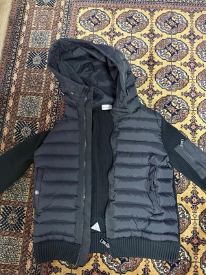 Svart pufferjacka från Moncler - Snygg svart pufferjacka från Moncler med stickade ärmar och huva. Jackan har dragkedja framtill, två fickor med dragkedja och Moncler-logga på ärmen. Perfekt för kyliga dagar och ger en sportig vibe.