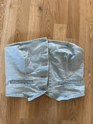 Ljusblå denim tubtopp - Trendig ljusblå tubtopp i denim med knappdetaljer framtill och smockad baksida för extra komfort. Toppen har två dekorativa fickor framtill och är perfekt för en cool och avslappnad look.