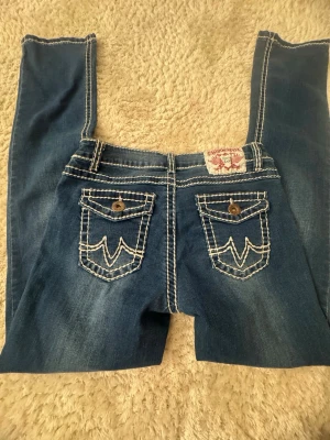 Blå Fashion Denim jeans med kontrastsömmar - Säljer ett par blå jeans från Fashion Denim med tydliga vita kontrastsömmar och dekorativa bakfickor med knappar. Jeansen har en klassisk femficksmodell och ett broderat märke bak i midjan. Materialet är denim i bomull.