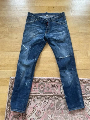 Mörkblå Dsquared2 ICON jeans herr - Snygga mörkblå jeans från Dsquared2 med ikonisk 'ICON'-patch bak. Jeansen har slitna detaljer, färgstänk och en smal passform. Klassisk femficksmodell med dragkedjegylf. Perfekta för en trendig streetwear-look.