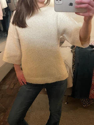 Krämvit stickad tröja med vida ärmar - Mysig krämvit stickad tröja med trekvartsvida ärmar och rund hals. Tröjan har en rak passform och ribbstickade muddar vid nederkant och ärmslut. Perfekt för lager-på-lager och enkel att styla till jeans eller kjol. Lite nopprig