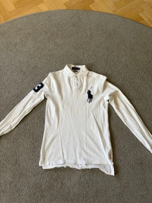 Vit långärmad skjorta från Polo Ralph Lauren - Vit långärmad skjorta från Polo Ralph Lauren med klassisk krage, knappar framtill och stor marinblå logga på bröstet. Snygg detalj med siffran 3 på ena ärmen. Tillverkad i mjuk bomull för en bekväm känsla.