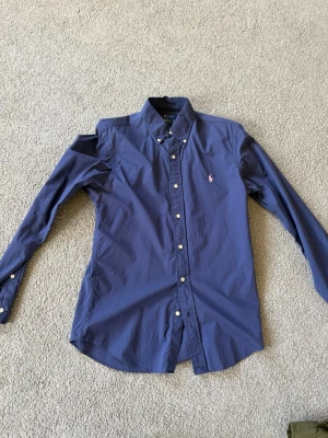 Blå skjorta från Ralph Lauren - Klassisk blå skjorta från Ralph Lauren med lång ärm och knappar framtill. Skjortan har en broderad rosa logga på bröstet och button-down krage. Perfekt för en stilren look.