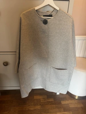 Ljusgrå ullkappa Zara - Stilren ljusgrå kappa med rak passform, stora framfickor och en dekorativ knapp vid halsen. Kappan är handgjord och har en enkel, modern design utan synliga sömmar eller detaljer. Perfekt för dig som gillar en clean och avslappnad look.