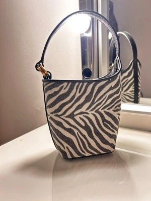 Mango handväska  - Snygg handväska med zebramönster i svart och vitt. Väskan har ett kort axelband med guldfärgad detalj. Köptes från mango förra sommaren, använder två gånger. Köptes för 520 säljer den för 399