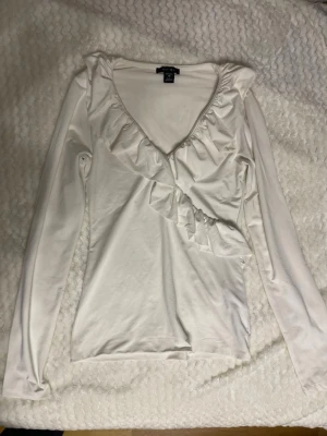 Vit långärmad topp med volang - Snygg vit långärmad topp med djup v-ringning och volangdetaljer framtill. Tillverkad i mjukt bomullsmaterial som känns skönt mot huden. Perfekt för att lyfta vilken outfit som helst med sin feminina touch.