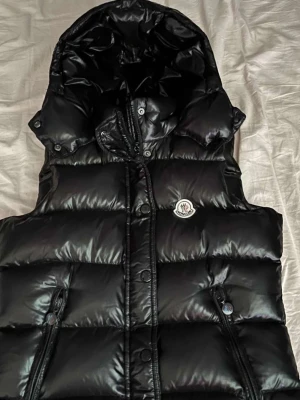 Svart dunväst från Moncler med huva - Snygg svart dunväst från Moncler med glansig finish och avtagbar huva. Västen har två dragkedjefickor fram och tryckknappar längs dragkedjan. Klassisk Moncler-logga på bröstet. Perfekt för lager på lager och streetwear.