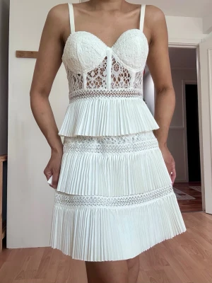 Student dress från ASOS Design - Superfin vit kortklänning från ASOS Design med spetsliv, plisserade volanger och smala axelband. Klänningen har genombruten spets upptill och flera lager plisserad kjol för en extra cool look. Perfekt för dig som vill sticka ut med detaljer och romantisk vibe.