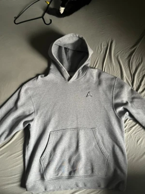 Grå Jordan hoodie med logga - Snygg grå hoodie från Jordan med klassisk Jumpman-logga broderad på bröstet. Hoodien har en stor känguruficka framtill och en rejäl huva. Tillverkad i mjukt och bekvämt material, perfekt för en avslappnad streetwear-look.