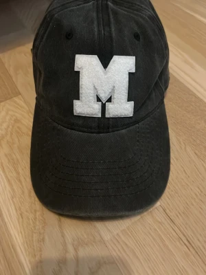 trendig keps  - Snygg svart keps med en stor vit M-bokstav framtill. Kepsen har böjd skärm. Perfekt accessoar för att lyfta din outfit. Använd endast en gång!💕