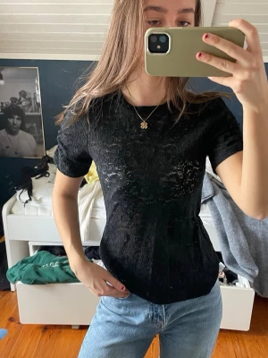 Svart spetsblus med korta ärmar - Snygg svart blus med spetsdetaljer och korta ärmar. Blusen har en feminin och elegant look med diskret mönster i spetsen. Perfekt att matcha med jeans eller kjol för en cool stil.