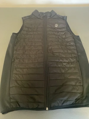 Svart dunväst från Jack & Jones - Stilren svart dunväst från Jack & Jones med quiltad framsida, hög krage och dragkedja hela vägen. Västen har två sidofickor med dragkedja och en diskret logga på bröstet. Perfekt för lager-på-lager och enkel att matcha med olika outfits.