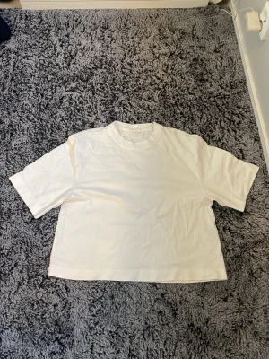 Vit croppad t-shirt - En enkel vit croppad t-shirt med rund hals och korta ärmar. Perfekt basplagg som passar till det mesta och är lätt att styla. Säljer pågrund av att jag gillar inte att den är så stor.