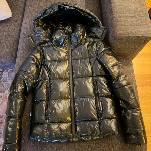 Grön pufferjacka från Khujo - Säljer en glansig grön pufferjacka från Khujo med stor huva och hög krage. Jackan har två dragkedjeförsedda fickor framtill och quiltad design. Perfekt för kalla dagar och riktigt snygg streetstil.