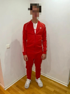 Adidas Orginals - SST TT Tracksuit  - Röd tracksuit från Adidas med vita ränder längs ärmar och ben samt Adidas logga på både tröjan och byxor. Setet består av en zip-jacka och matchande byxor i sportig, stretchig polyester. Perfekt för en avslappnad och sportig stil. Storlek S på tröjan och storlek M på byxan. Köptes för 2 år sen retail 1599 kr. 