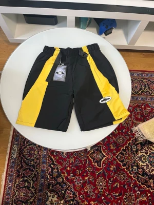 Svarta och gula shorts med t shirt som matchar  - Snygga svarta shorts med gula paneler. De har elastisk midja och sportig look med logga på ena benet. Perfekta för sommaren och streetwear-stil. 