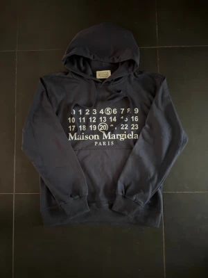 Maison Margiela svart hoodie - Snygg svart hoodie från Maison Margiela med ikoniskt tryck framtill i vitt med siffror och logga. Klassisk huva med dragsko och stor magficka. Perfekt för dig som gillar streetwear och vill sticka ut med en exklusiv look. Passar S/M