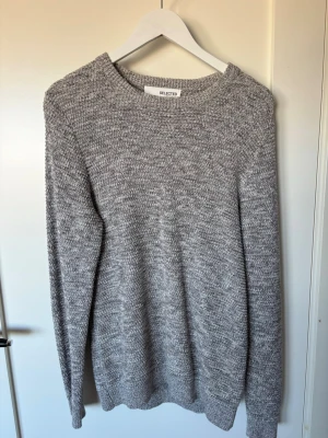 Grå stickad tröja från Selected - En stilren gråmelerad stickad tröja från Selected. Tröjan har rund halsringning, långa ärmar och ribbade muddar vid ärmslut och nederkant. Perfekt för lager-på-lager och enkel att matcha med jeans eller chinos. Inte använt pga spontanköp som jag senare märkte att jag inte gillade. 