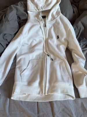 Vit hoodie från Polo Ralph Lauren - Snygg vit hoodie från Polo Ralph Lauren med dragkedja framtill och klassisk logga broderad på bröstet. Hoodien har kängurufickor, ribbade muddar och justerbar huva med snören. Perfekt för en clean och stilren look.