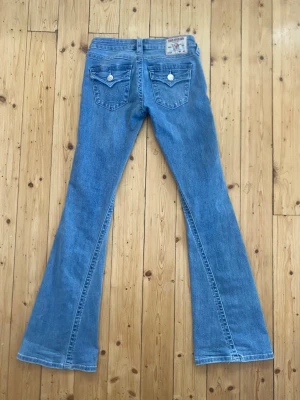 True Religion ljusblå flare jeans - Säljer ett par ljusblå low rise flare jeans från True Religion, modell Joey. Jeansen har snygga fickor bak med lock och vita knappar, samt klassiska sömmar och detaljer. Perfekta för dig som gillar retrovibbar och vill ha ett par riktigt schyssta jeans.